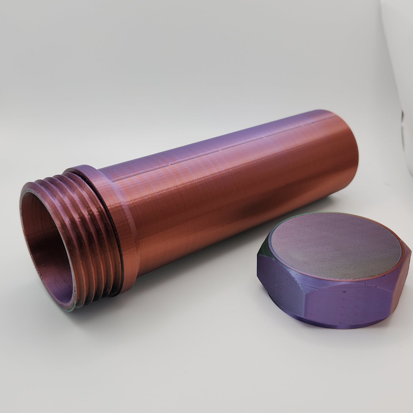 The cylindrical container is shown lying on its side with the hexagonal cap removed. The container displays threading inside, indicating it is designed to be securely closed. The gradient color transition from purple to green to red is clearly visible on both the container and the cap.