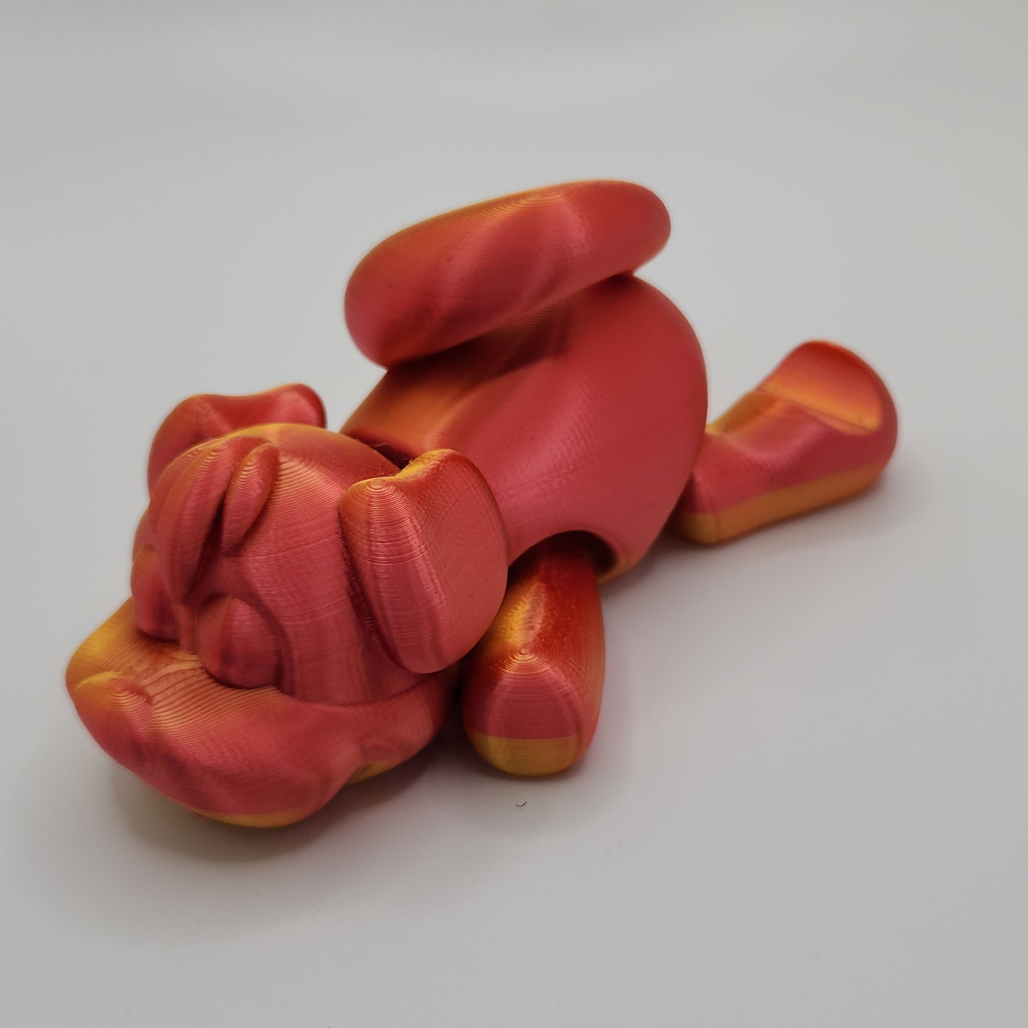 The side view of a 3D-printed dog figurine in red and gold hues. The dog is in a playful pose, laying on its stomach with its front paws stretched out and head resting on the ground. The glossy finish highlights the smooth and shiny surface of the print.