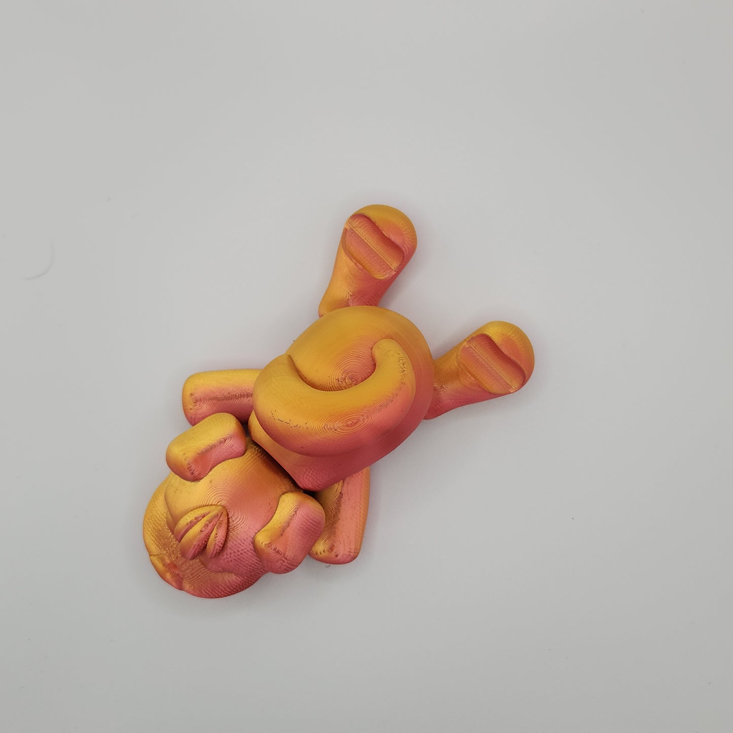 A top-down view of a 3D-printed dog figurine in red and gold shades. The dog is laying flat on its stomach with its legs spread out, showcasing the smooth and shiny surface of the print. The curled tail and the dog's relaxed pose are clearly visible from this angle.