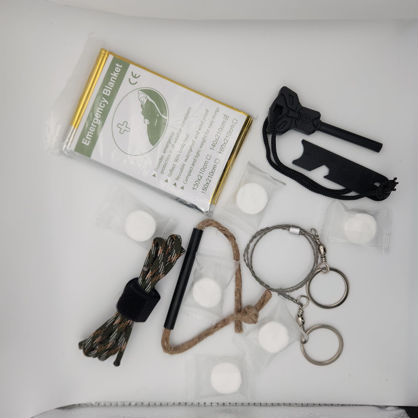 A close-up view of the hiking supplies. The focus is on the same assortment of items: a camo paracord, black firestarter with a lanyard, individually wrapped compressed towels, a metal wire saw with rings, an emergency blanket in its package, and a twine loop with a black metal tube.