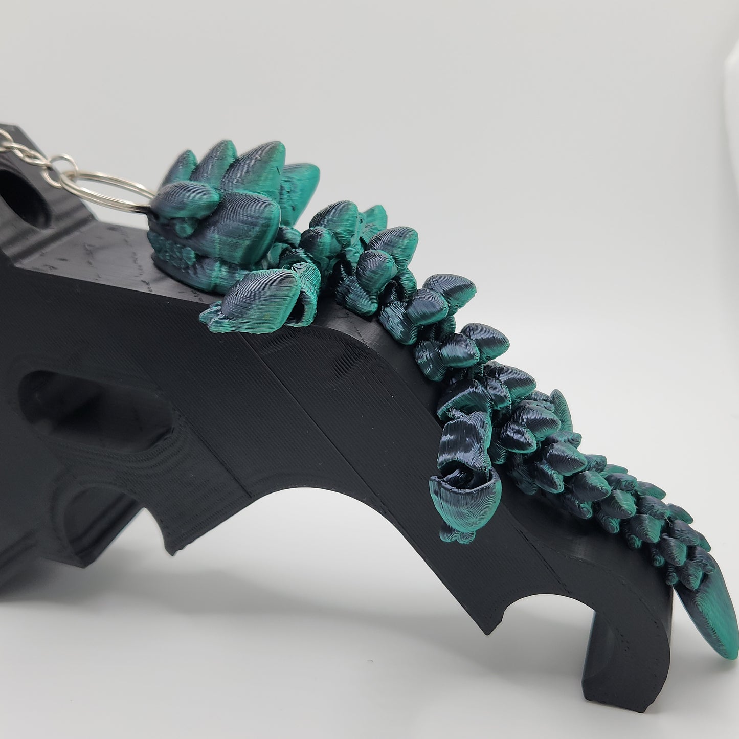 A 3D printed articulated dragon keychain in black and teal, shown climbing a black object with intricate scale details.