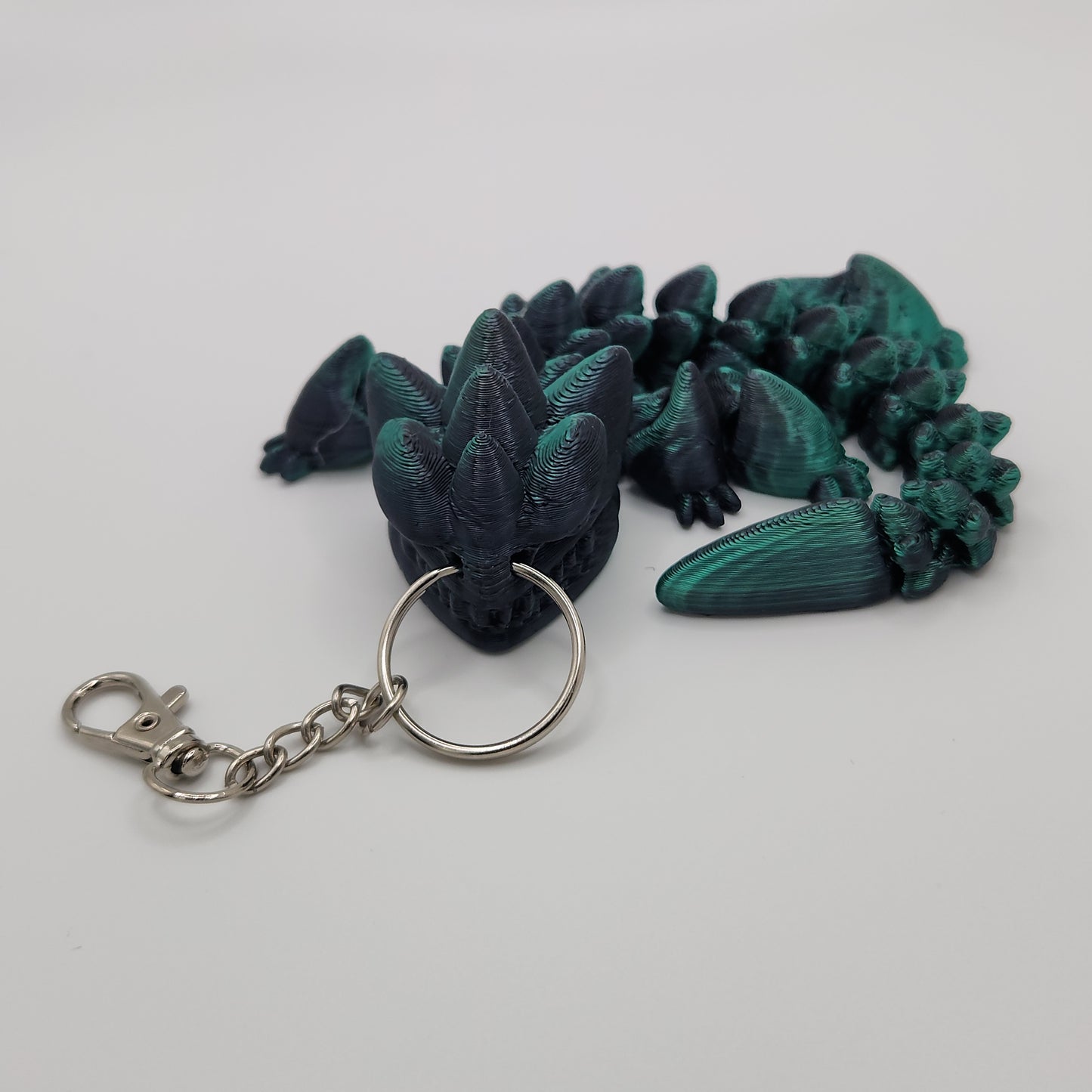 The articulated dragon keychain curled up on a white background, highlighting its black and teal scales and attached keyring.