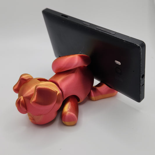 A 3D-printed dog figurine in a blend of red and gold colors is holding a black phone in its back. The dog is designed with exaggerated features, including large ears and paws, giving it a cartoonish appearance. The phone is securely placed between the dog's back and the ground.