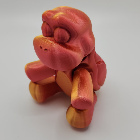 A front view of a 3D-printed turtle figurine in red and gold colors. The turtle has a detailed shell with segment patterns and a cartoonish face with large, closed eyes and a wide mouth. The figurine has a glossy finish, highlighting the smooth texture.